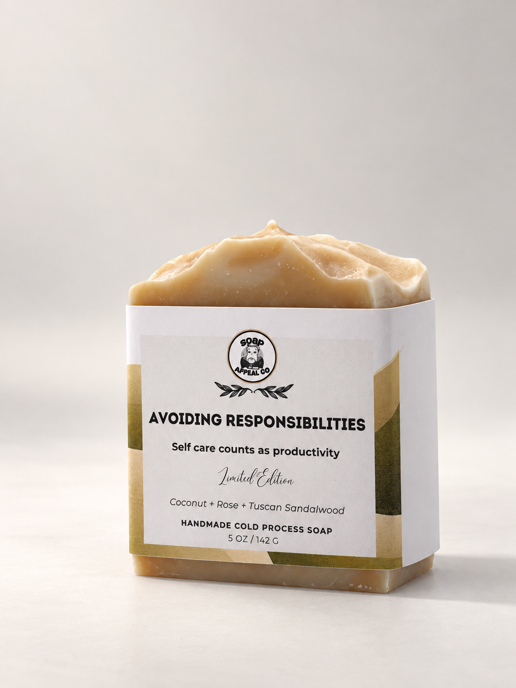 A handmade cold process soap bar wrapped in a white paper sleeve with black text, set against a clean, light gray background. The soap has a natural, textured tan color.
