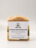 A single bar of handmade cold process soap in a square glass container with a textured, swirled top, wrapped in a white paper label. The soap has a warm beige/cream colour.