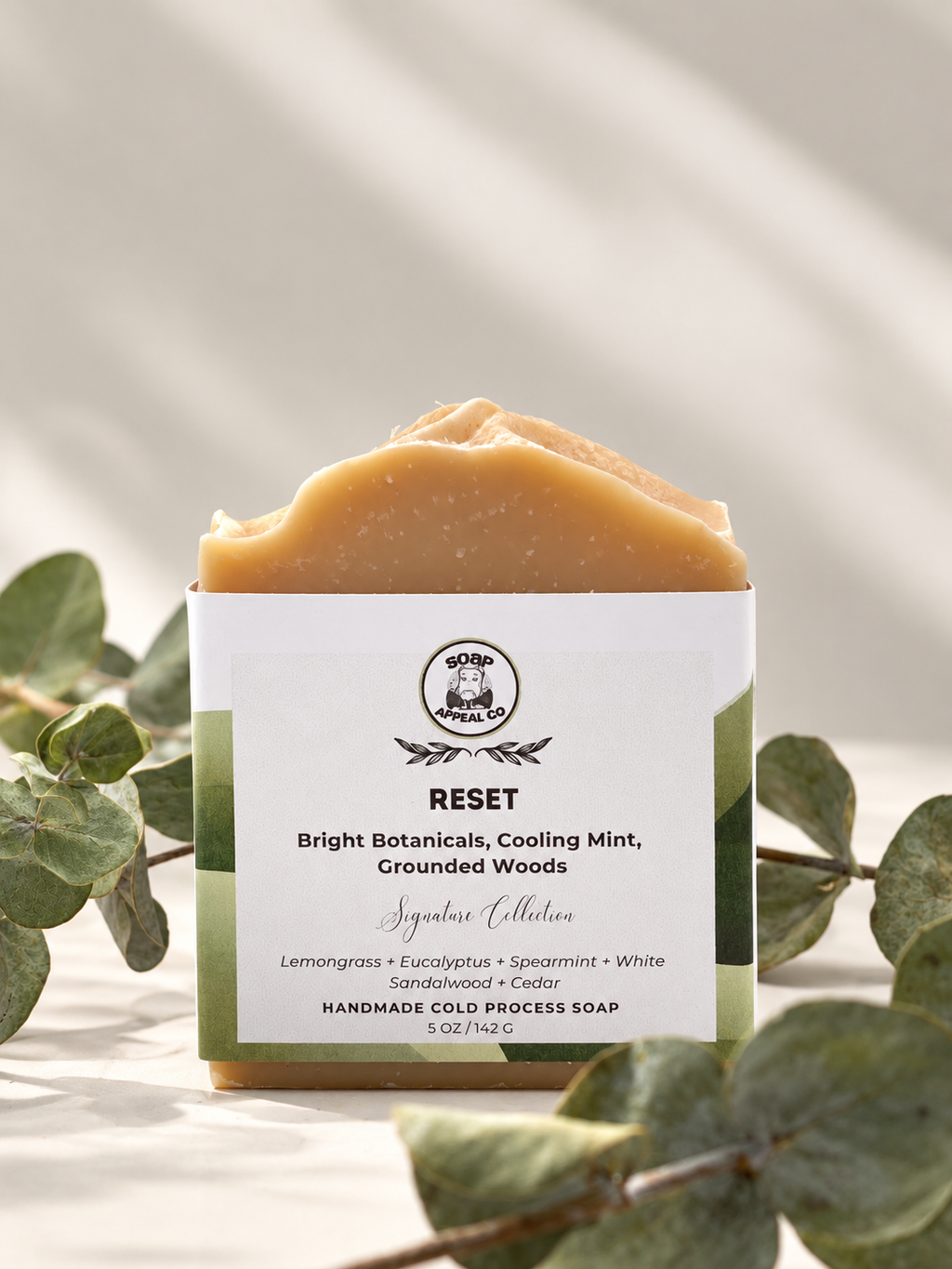 A single bar of handmade cold process soap labeled 'RESET' by Soap appeal, styled with green eucalyptus leaves on a light beige background with soft shadows.