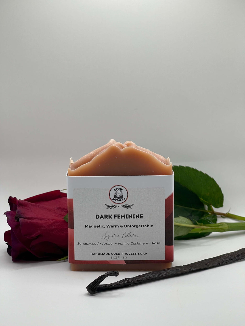 Bar of soap with a white label on a light gray background, surrounded by a rose and vanilla bean.