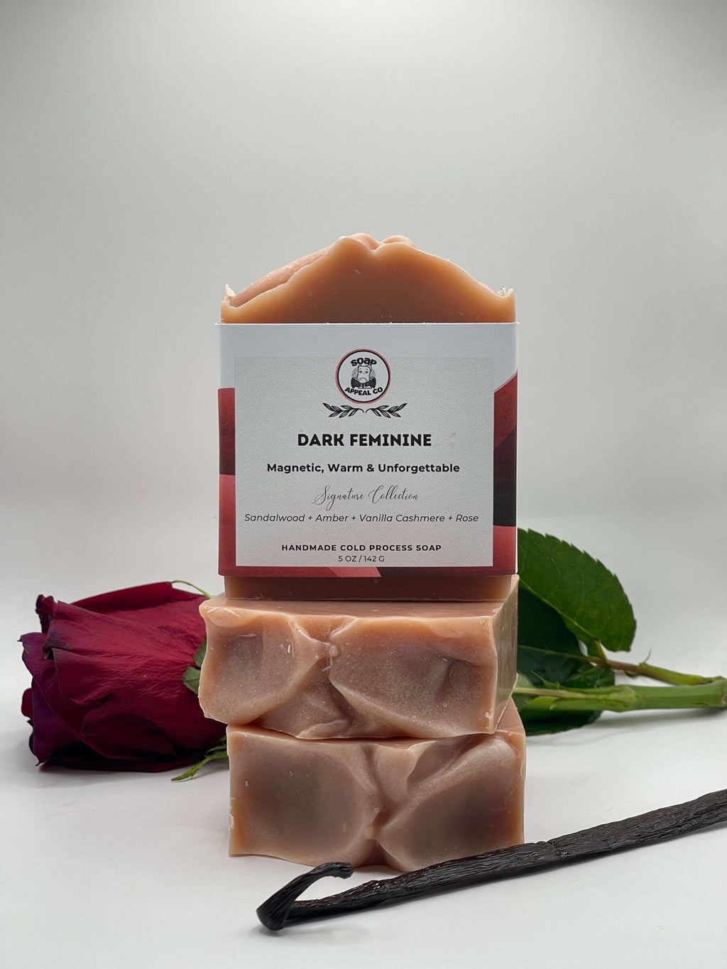 Stack of soap bars with a 'Dark Feminine' label, rose, and vanilla bean on a white background
