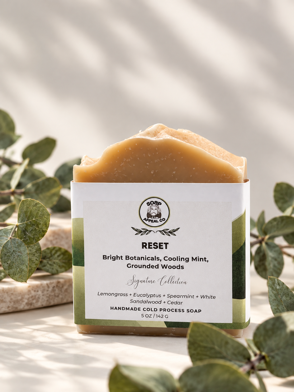 A rectangular bar of handmade cold process soap in a square container with a white label, styled with green eucalyptus leaves on a light surface with soft shadows. The soap bar is a warm tan/golden color.