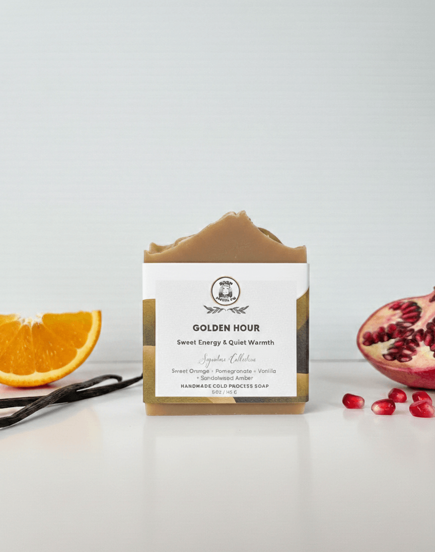 Bar of soap with a label on a white surface, surrounded by an orange, pomegranate, and vanilla bean.