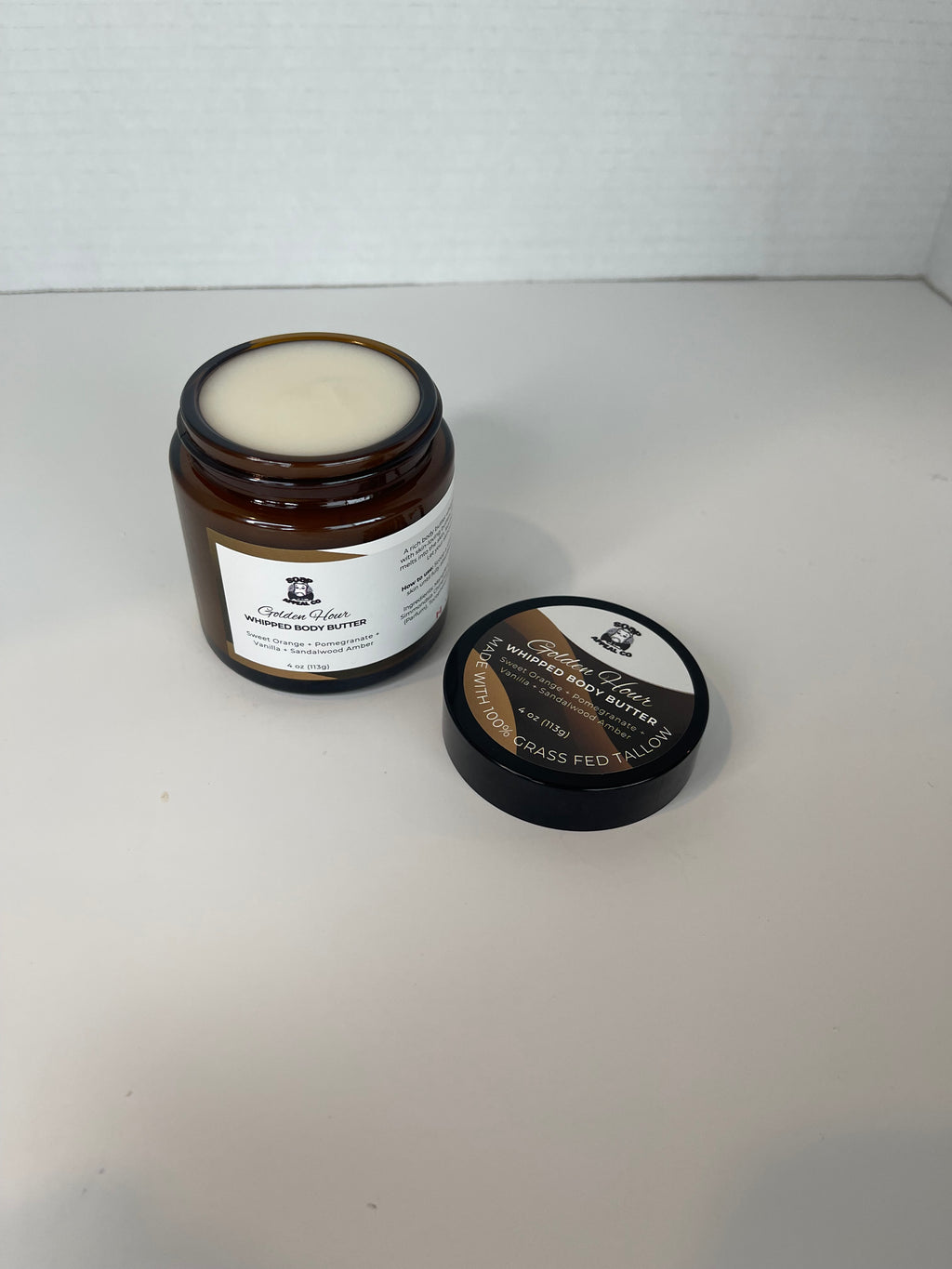 Lid off the jar with labels on a white surface to show the body butter in the jar