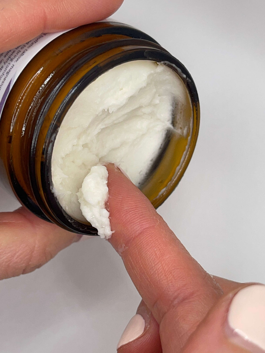Hand holding a jar of cream with a finger applying some to the skin.