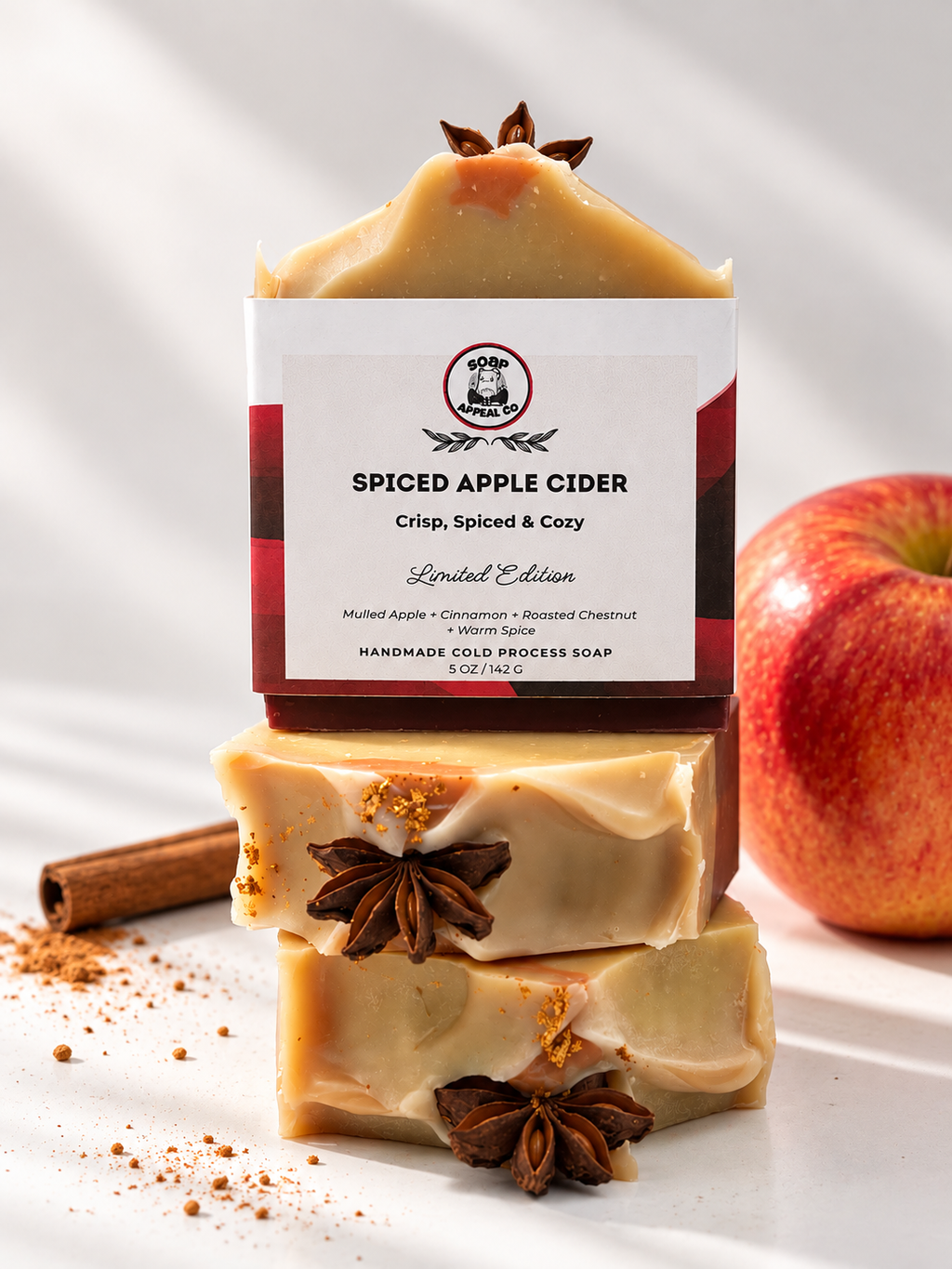 Three stacked bars of Spiced Apple Cider handcrafted cold process soap on a white surface, styled with a cinnamon stick, apple slices, and star anise. The packaging label is clearly visible in the center.