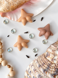 Three starfish-shaped soaps in natural beige and peach tones, styled on a white surface with seashells, glass rhinestones, and wooden beads.