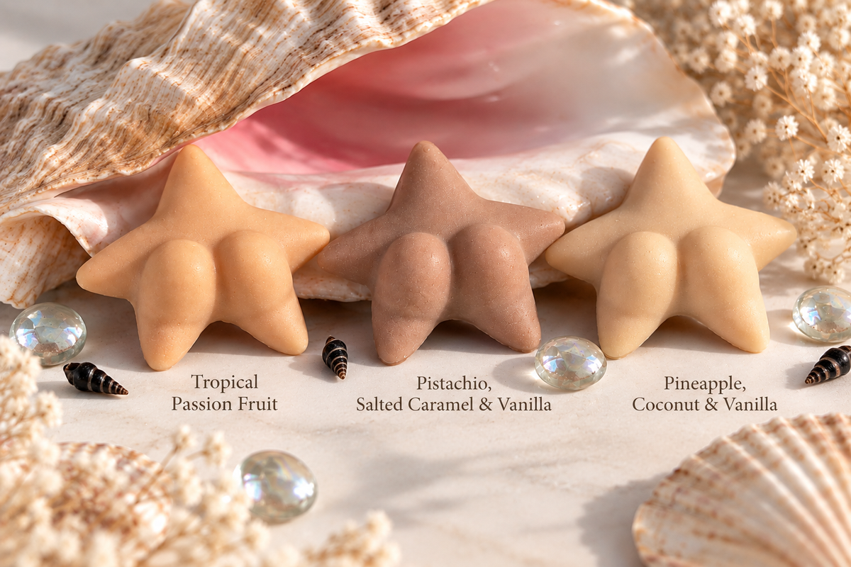Three star-shaped soap bars in tropical shades labeled Tropical Passion Fruit, Pistachio, and Pineapple, Coconut & Vanilla, styled with seashells and sea glass on a light surface.