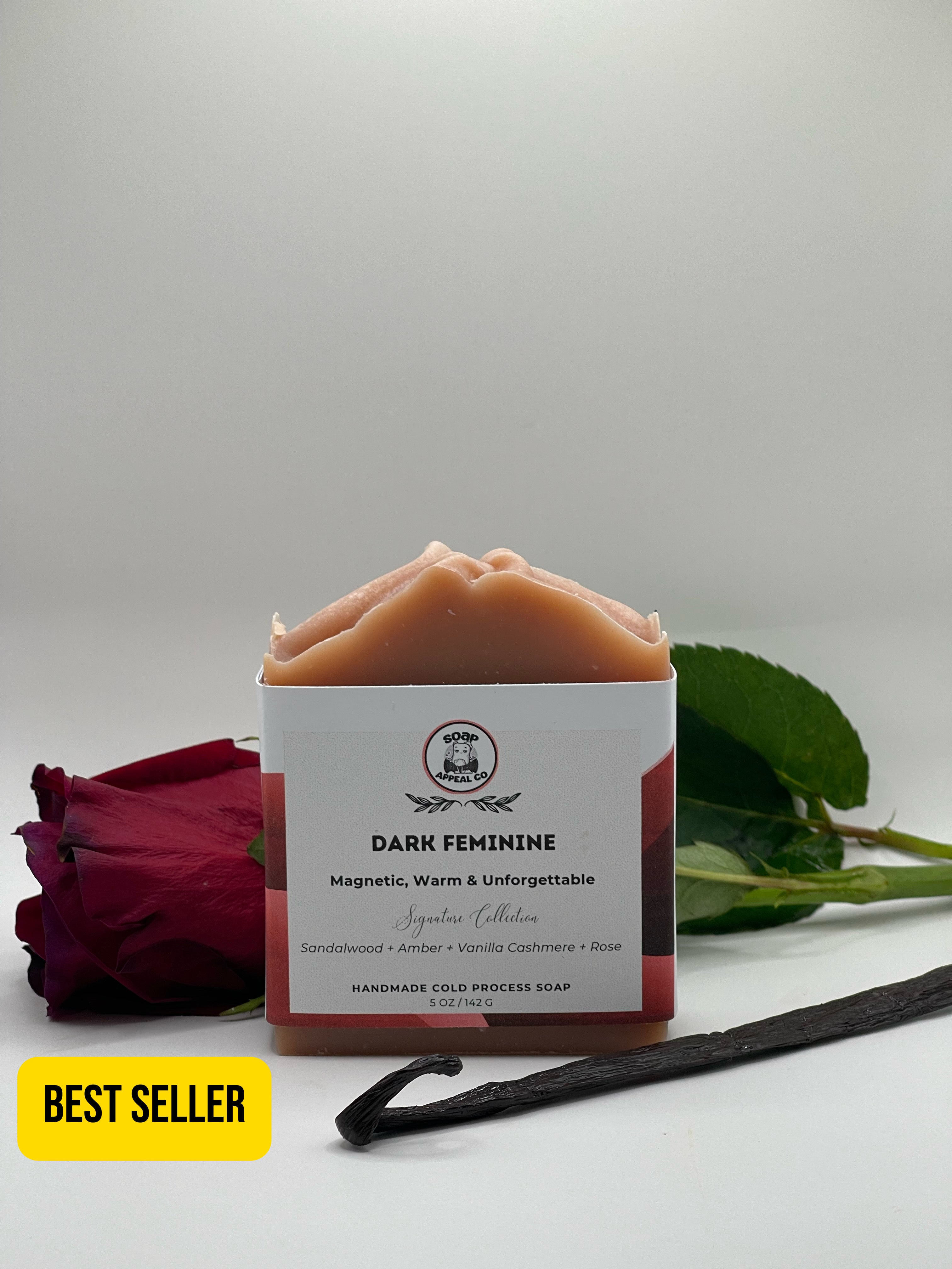 A square bar of handmade cold process soap labeled 'DARK FEMININE' from the Signature Collection, styled with a red rose, a vanilla bean, and a yellow 'BEST SELLER' badge on a light gray background.