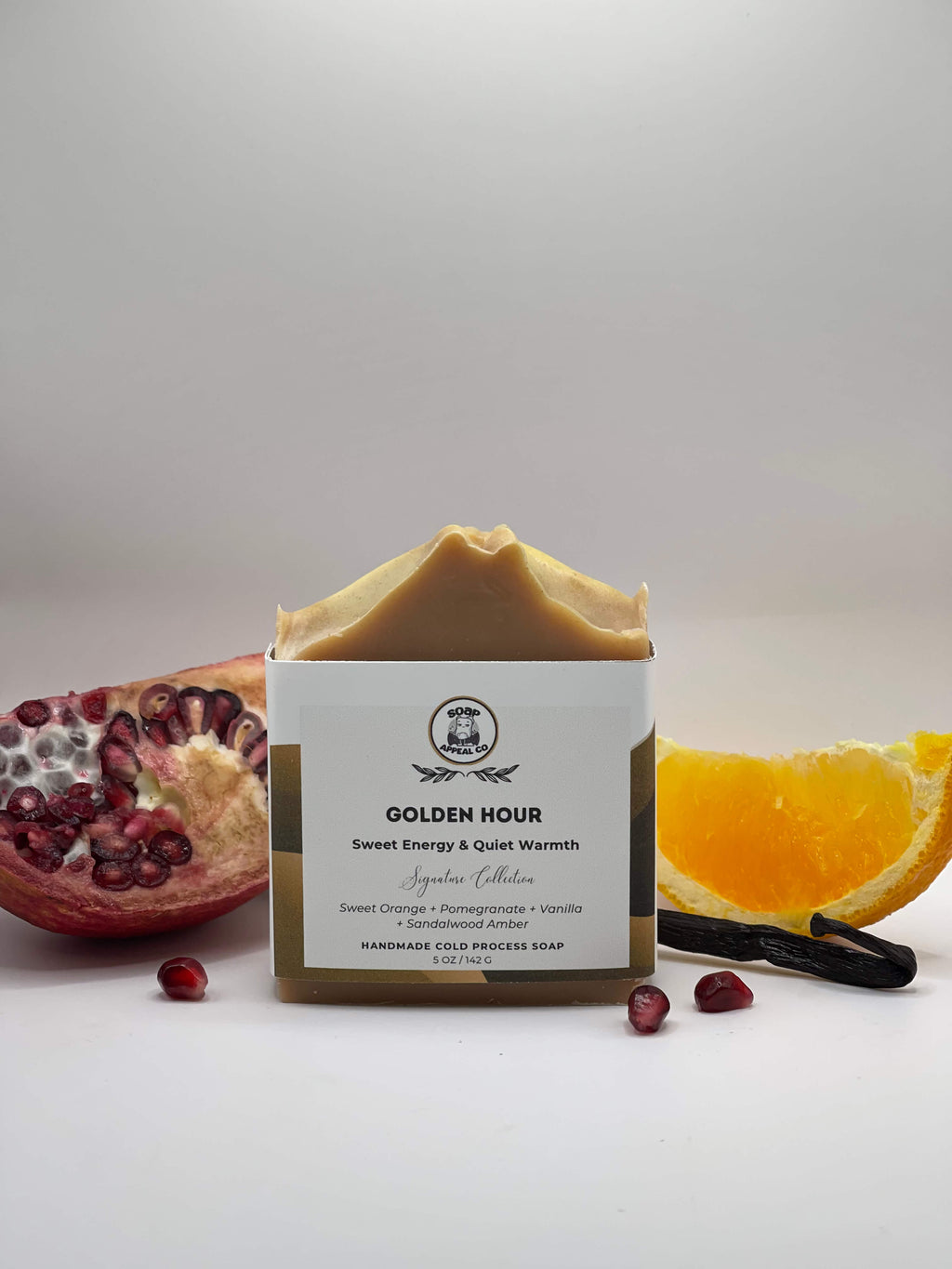 Bar of soap with packaging labeled 'Golden Hour' next to a pomegranate, orange, and vanilla bean on a light background
