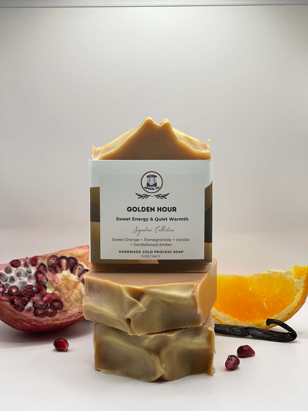 Bar of soap with a label titled 'Golden Hour' surrounded by pomegranate, orange, and vanilla bean on a white background.